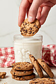 A hand is captured placing a chocolate chip cookie atop a full glass of milk, with a stack of cookies and a checkered napkin beside it