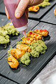 Anonymous chef carefully applies a red sauce to crispy fried snacks on a contemporary slate tray, garnished with green moss