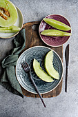 Top view of freshly sliced organic mini melons arranged elegantly on decorative plates, accompanied by a dark metal fork and a rustic wooden board, ready for a refreshing lunch.