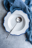 An elegant white plate with blue edges and a silver spoon, accompanied by a denim blue napkin on a textured surface