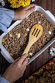 Top view of a cropped unrecognizable person cooking an homemade meat pie with pieces of potatoes, stirred with a wooden spoon in a white baking dish, adjacent to flowers and a decorative cloth.