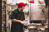 A professional chef, sporting a red turban and black uniform, attentively cooks in a bustling commercial kitchen environment