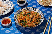 Top view of a traditional Chinese cuisine setup on a vibrant blue tablecloth. The scene includes a plate of stir-fried noodles with shrimp and vegetables, alongside dumplings, and dishes of soy and chili sauces