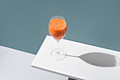 A wine glass filled with fresh carrot juice balances on a white edge, casting a long shadow on a muted teal background, showcasing a blend of elegance and minimalism.