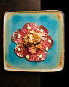 Artistic presentation of beef carpaccio adorned with dollops of sauce, edible flowers, and crispy toppings, elegantly served on a rustic blue plate against a dark background