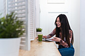 Smiling female sitting at counter with cocktail in bar and writing in notepad while looking at camera