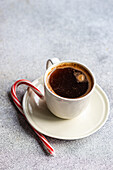 A cozy morning scene featuring a white mug of hot coffee paired with a red and white candy cane on a speckled grey surface.