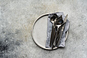 Top view of modern minimalist tableware set up with a ceramic plate, neatly folded grey napkin, and assorted silver cutlery on a concrete backdrop