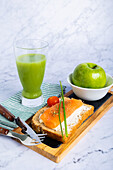 Nutritious meal featuring a glass of cucumber and apple juice, a green apple, fresh salmon on whole grain toast decorated with herbs and cherry tomato, set on a marble table. Perfect for a wholesome breakfast or light meal.