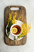 From above of inviting cup of tea with a lemon slice on a wooden board, adorned with fragrant Mimosa sprigs, captures the essence of a leisurely morning