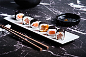 Assorted sushi rolls served on a white plate with soy sauce and chopsticks on a dark marble background.