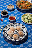 Top view of a traditional Chinese food arrangement featuring shrimp noodles, dumplings, sliced cucumbers, and sauces on a blue patterned tablecloth, illuminated by natural sunlight