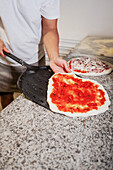 Ein gesichtsloser Pizzabäcker verteilt Tomatensoße auf dem Pizzateig und hat eine rohe Pizza mit Schaufel im Blick.