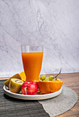 Vibrant selection of fresh fruit including kiwi, orange, apple, and grapes, alongside a glass of orange juice. All items are neatly arranged on a circular plate, set against a marble background with a rustic wood surface.