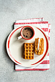 A delightful top-down view of homemade waffles filled with vanilla marshmallow, alongside a cup of coffee, all served on a plate with red stripes resting on a patterned napkin