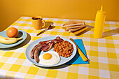 Yellow coloured Tablecloth with a delicious full English Breakfast, egg, beans, sausages and bacon, some toasted bread slices, fruit and a cup of coffee