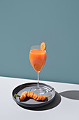 A glass of carrot juice with a slice on the rim and a whole carrot on a plate, set against a teal background with shadow.