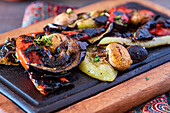 A vibrant assortment of grilled vegetables including bell peppers, mushrooms, and eggplants presented on a dark wooden serving board, seasoned with fresh herbs