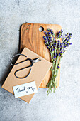 A rustic wooden cutting board holds a bouquet of fresh lavender flowers next to scissors and a thank you card, creating a welcoming summer table setting