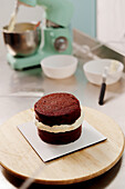 A freshly baked layered cake with creamy frosting on a wooden plate, with a mixer and bowls in the background, depicting a home baking scene