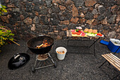 A charcoal grill actively burning, next to a table lined with sliced vegetables, meats, and buns for a barbecue setup outdoors, against a stone wall backdrop.