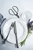 A picturesque summer table setting featuring white plates, modern cutlery, a glass tumbler, and fresh lavender flowers, all arranged on a light grey surface with a hint of warmth and sophistication