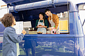 Cheerful friends serve a happy customer from their vibrant blue food truck, reflecting a friendly street food business vibe