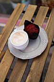 Ein frischer Muffin aus rotem Samt und ein heißer Cappuccino auf einem Vintage-Teller, der auf einem Holztisch für eine entspannte Kaffeepause steht