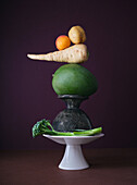 An imaginative arrangement of vegetables, featuring a parsnip, potato, tomato, and green mango balanced atop a rustic stone podium, all set against a deep purple backdrop