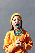 A young individual in an orange sweatshirt and yellow beanie savors the taste of a large, vibrant swirl lollipop against a plain background