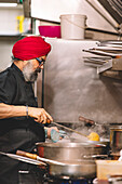 A Sikh chef, distinguished by his red turban, attentively cooks in a bustling commercial kitchen with steam rising from the pots