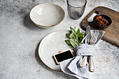 An elegantly simple summer table setting featuring a neutral palette with fresh mint leaves as a central theme The image includes beautifully textured ceramic dishes, a glass, silverware wrapped in a grey napkin