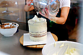 An individual meticulously adds a layer of frosting to a towering, multi-layered cake using a metal spatula, with bowls of ingredients nearby