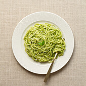 Overhead shot of a white plate filled with spaghetti coated in vibrant green pesto sauce, served on a beige linen tablecloth.