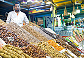 A vendor, appearing Middle-Eastern, looks at the camera while standing behind a colorful display of dates, nuts, and dried fruits at a lively market stall in Marrakesh.