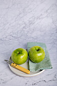 Two vibrant green apples served on a light-colored plate with a knife, resting on a striped napkin on a marble background, illustrating a healthy, wholesome diet.