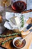 A wedge of Brie cheese on a wooden board, accented with rosemary, next to honey and a glass of white wine.