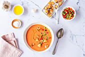 Top view of a traditional Spanish Tomato Gazpacho Soup served with croutons and diced vegetables alongside olive oil and garlic, arranged on a marble surface.