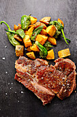 Juicy, cooked steak with coarse salt paired with golden roasted sweet potatoes and blistered green peppers on a dark slate surface.