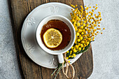 Top view of inviting cup of tea with a bright lemon slice on an ornate saucer, accompanied by a delicate Mimosa flower bundle, presented on a wooden board with a textured backdrop