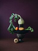 Fresh vegetables arranged with a vintage juicer, featuring broccoli, turnips, an orange, and potatoes against a rich maroon background, portraying a mix of rustic and modern kitchen themes