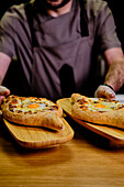 Ein Koch, teilweise sichtbar, serviert frisch gebackene georgische Khachapuri, ein mit Käse gefülltes Brot mit einem Spiegelei in der Mitte, präsentiert auf Holzbrettern vor einem dunklen Hintergrund