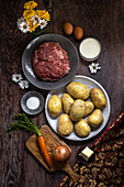 Top view of fresh ingredients for preparing a homemade meat pie, featuring ground beef, potatoes, butter, milk, eggs, onion, carrot, and salt, all neatly arranged on a rustic wooden table.