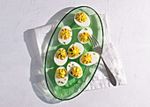 Top view of a fresh preparation of deviled eggs, neatly arranged on a vibrant green glass plate complemented by a silver fork, set against a textured white background.