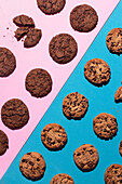 A flat lay photograph featuring an assortment of chocolate chip and double chocolate cookies arranged on a dual background of pink and blue. One cookie on the pink side is broken, adding a touch of visual interest