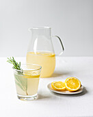 A pitcher full of refreshing lemonade beside a filled glass garnished with rosemary, with halved lemons on a saucer against a white background