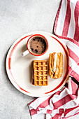 Goldene hausgemachte Waffeln mit Vanille-Marshmallow-Füllung und eine Tasse heißer Kaffee auf einem rustikalen Teller mit gestreiftem Handtuch