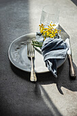 Top view of fresh outdoor table setting featuring a white embossed plate, silver cutlery, a blue linen napkin, and a sprig of bright yellow mimosa flowers