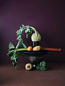 A harmoniously balanced still life featuring a kohlrabi with attached leaves, resting inside a vintage metal bowl, surrounded by an assortment of vegetables including broccoli, potato