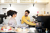 Friendly barista in a yellow apron chats with colleague by the espresso machine with cakes on display.